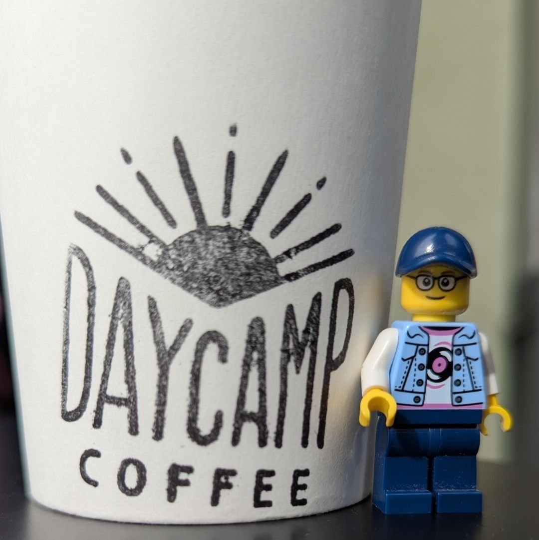 A close-up image of a coffee cup featuring a logo that reads DAYCAMP COFFEE with a sun design above it. Next to the cup, there is a LEGO figure wearing a blue cap, glasses, and a camera around its neck.