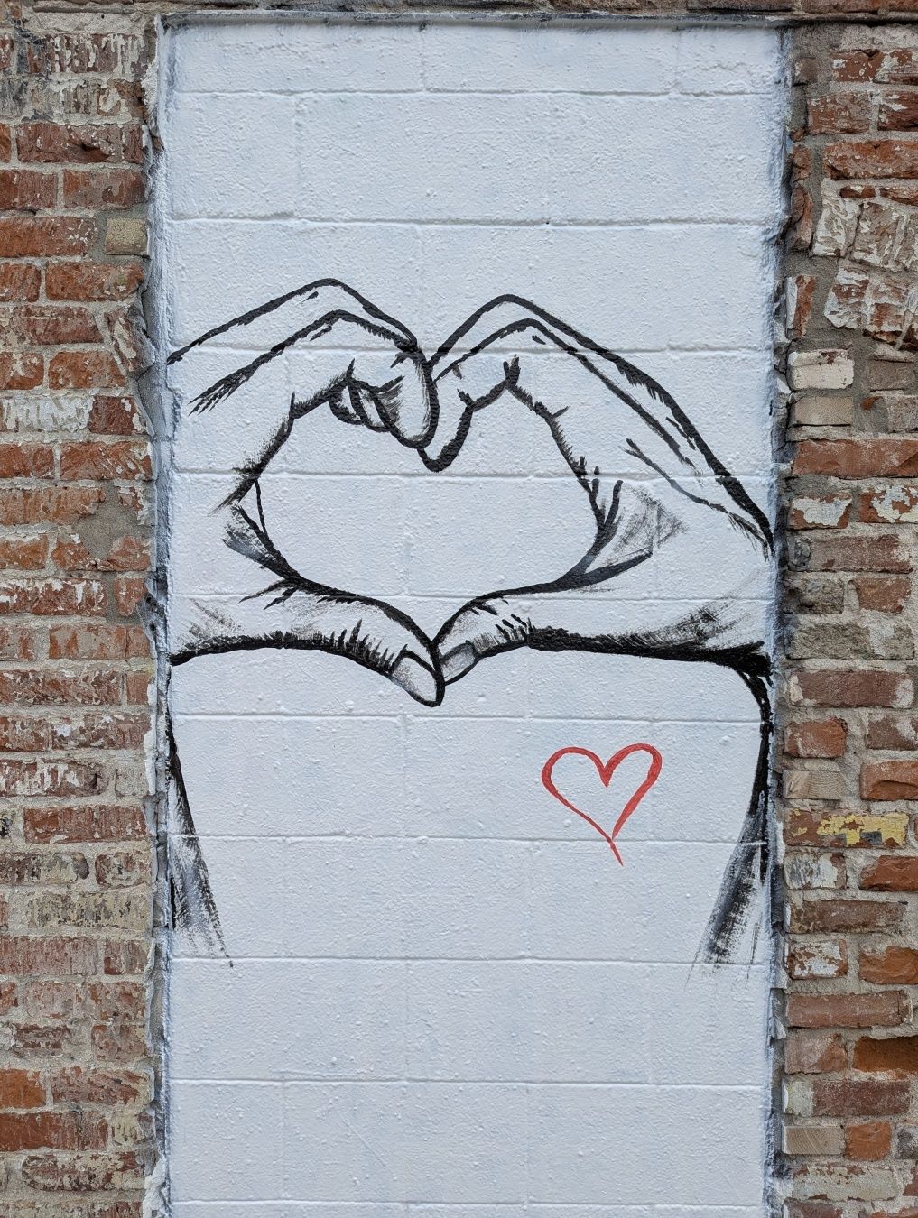 A mural depicting two hands forming a heart shape against a textured brick wall. Below the hands, there is a small red heart drawn. The artwork uses a monochromatic style for the hands, contrasting with the colorful brick background.