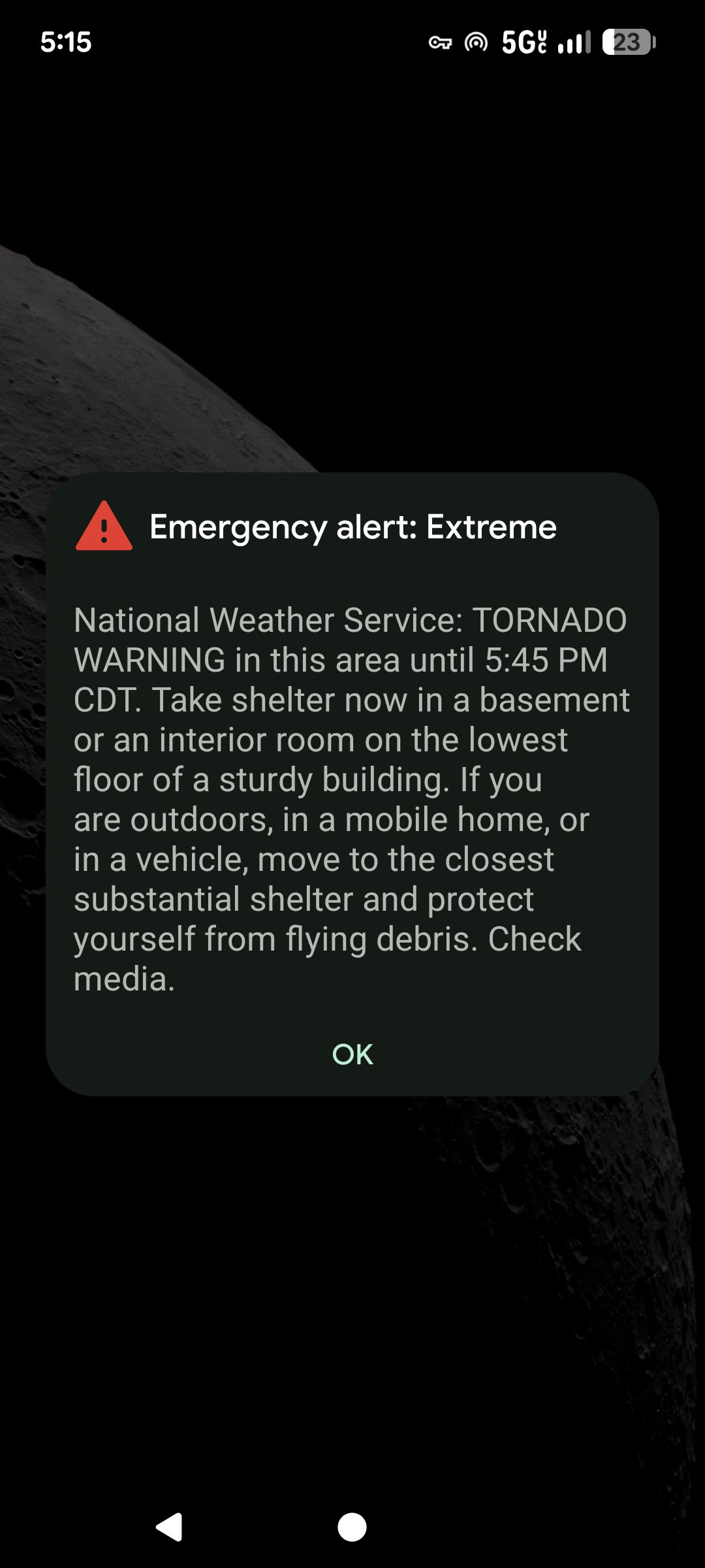 Screenshot of a phone showing an emergency alert. A dark-themed notification box with a red triangle icon reads: “Emergency alert: Extreme.” The alert text says: “National Weather Service: TORNADO WARNING in this area until 5:45 PM CDT. Take shelter now in a basement or an interior room on the lowest floor of a sturdy building. If you are outdoors, in a mobile home, or in a vehicle, move to the closest substantial shelter and protect yourself from flying debris. Check media.” A single “OK” button appears below the message. The phone status bar and a partial moon background are visible.